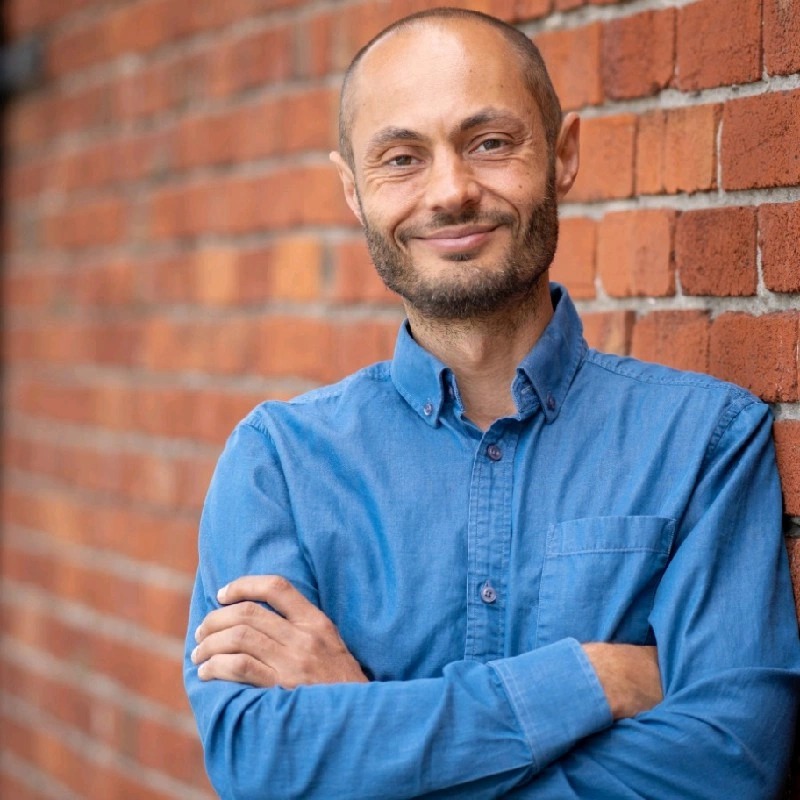 Antony Evans, smiling, arms folded, in a blue shirt against a red brick wall.